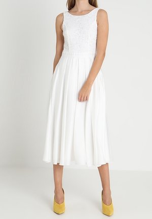 Kvinde iført ærmeløs hvid blondekjole med plisseret midi-nederdel og gule højhælede sko, stående mod hvid baggrund.