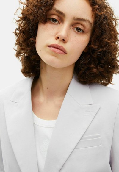 Blazer grigio chiaro con revers a punta, taschino sul petto singolo e tessuto texturizzato. Indossato sopra una maglia bianca con dettagli di design discreti.
