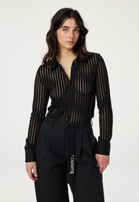 Black button-up shirt with sheer striped fabric, long sleeves, and cuffs featuring button details. Worn with high-waisted black trousers.