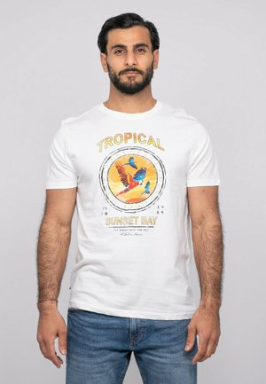 Homme aux cheveux foncés et barbe, portant un t-shirt blanc avec des oiseaux tropicaux colorés et le texte « Tropical Sunset Bay », debout devant un fond uni.