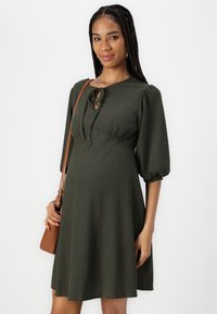 Grønn, knelang kjole med puffærmer og knytet hals. Laget av teksturert stoff. Kombinert med en brun skulderveske.