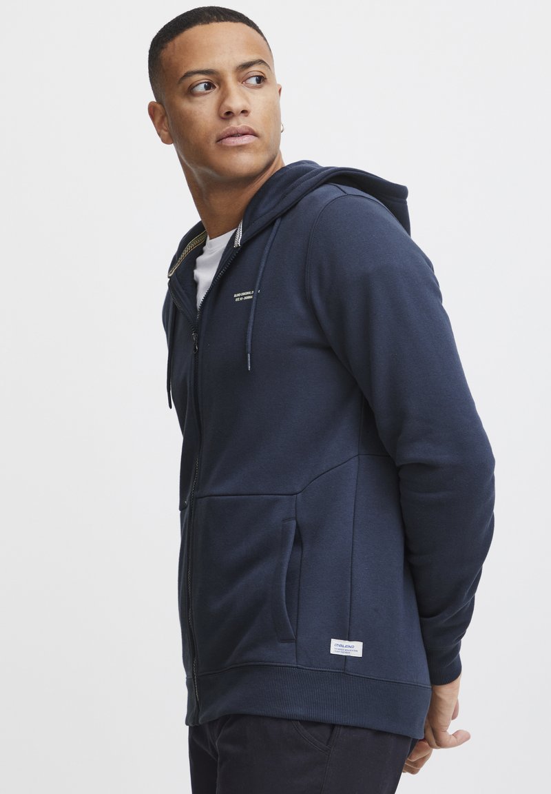 Marineblå zip-up hettegenser med snorsnor i hetten, lommer foran, ribbestrikkede mansjetter og logomerke i kanten, laget av et mykt, strukturert stoff.