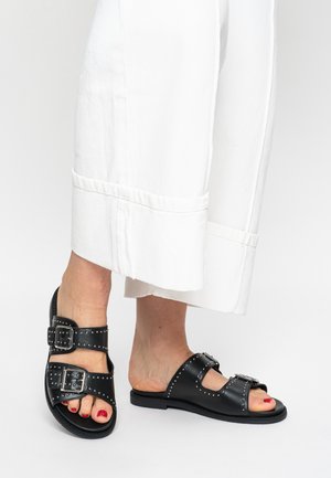 Black leather sandals with two wide straps, silver buckle accents, and studs, paired with white wide-leg pants.