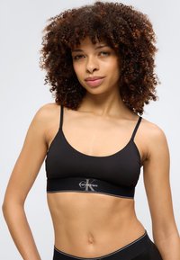 Νεαρή γυναίκα με σγουρά μαλλιά που φοράει ένα μαύρο bralette Calvin Klein και μαύρα κάτω μέρη, κοιτάζοντας προς τα εμπρός με ένα ελαφρύ χαμόγελο.