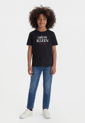 Barn med krøllet hår står iført sort Calvin Klein T-shirt, blå jeans, sorte sokker og hvide sneakers mod en hvid baggrund.