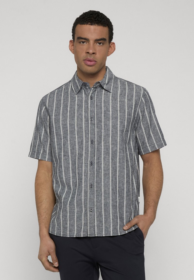 Homme portant une chemise grise à manches courtes, boutonnée, avec des rayures verticales blanches, debout avec une main dans la poche devant un fond uni.