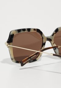 Koraliniai saulės akiniai su rudo atspalvio lęšiais, auksiniais akcentais ir aštuonkampiu rėmu. "Michael Kors" išgraviruota ant šventės.