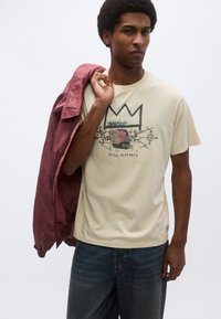 Μπεζ βαμβακερό T-shirt με γραφικό τύπωμα που απεικονίζει μια κορώνα και αφηρημένο σχέδιο. Το μοντέλο κρατά ένα μπορντό σακάκι στον ώμο, φορά τζιν παντελόνι.