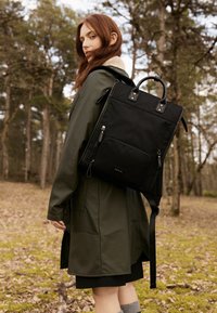 Black backpack with top handles and adjustable straps, featuring zippers, a front pocket, and a smooth texture, set in a forested area.