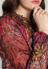 Blusa trasparente con motivo floreale in tonalità di borgogna, rosa e oro. Collo arricciato e polsini elasticizzati aggiungono texture e dettagli.