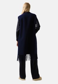 Námořnický modrý dlouhý vest, vyrobený z měkké látky, s třásňovým lemem. Nosený přes tmavý outfit, ukazuje bezramenný, volný střih.