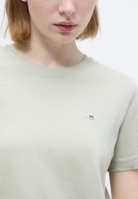 Κοντινό πλάνο ενός ατόμου που φοράει ένα ανοιχτό πράσινο T-shirt με ένα μικρό κεντημένο λογότυπο ασπίδας στο αριστερό στήθος.