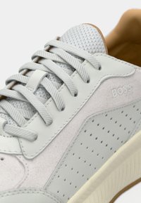 Ανοιχτό γκρι αθλητικό παπούτσι με γλώσσα από υφή διχτυού, επίπεδα κορδόνια, διάτρητη πλευρική επιφάνεια, λεπτομέρειες από σουέντ και ανάγλυφο λογότυπο "BOSS" κοντά στην φτέρνα.