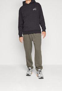 Felpa nera con cappuccio e tasca frontale, logo sul petto, abbinata a pantaloni della tuta verde oliva e scarpe da ginnastica grigie con dettagli arancioni.