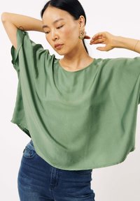 Groene blouse met korte mouwen gemaakt van een gladde stof; relaxte pasvorm met een ronde halslijn en subtiele glans, gecombineerd met blauwe denim jeans.