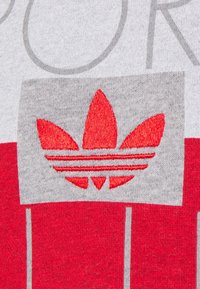 Červená a šedá mikina s velkým vyšitým logem, které zobrazuje tři listy a tři horizontální pruhy. Měkká textura, ležérní design.