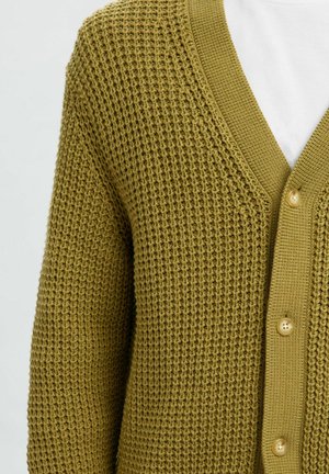 Olijf groene chunky gebreide cardigan met V-hals, knoopsluiting en wafelstructuur, gedragen over een wit shirt.