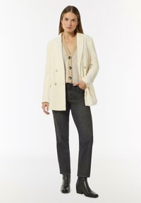 Doppelreihige cremefarbene Blazer mit strukturiertem Muster, kombiniert mit einem beigefarbenen Stricktop und dunklen, maßgeschneiderten Hosen, abgerundet mit schwarzen Ankle Boots.
