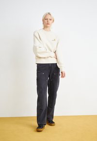Beige sweatshirt med ribbede manchetter og kant, logo detalje på brystet; mørkeblå vide cargo bukser med forlommer og guld accenter.