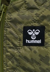 Grön quiltad jacka i tyg med ett subtilt mönster, med en svart etikett som visar "hummel"-logotypen på övre högra hörnet. Dragkedja på vänster sida.