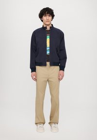 Giacca bomber navy con polsini e colletto a coste, chiusura con zip e due tasche frontali; indossata sopra una maglietta grafica multicolore e pantaloni beige.