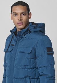Chaqueta de plumón azul con capucha, que presenta un cierre frontal con cremallera, dos bolsillos en el pecho y un tejido texturizado. Parche de logo de goma en la manga.