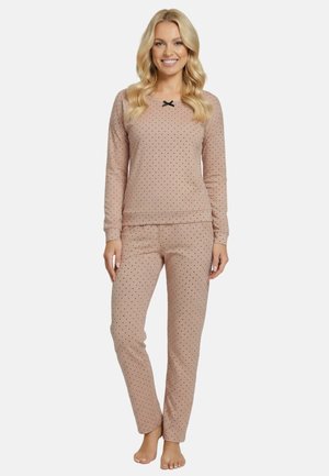 Kvinde smilende, iført beige polkaprikket langærmet top og matchende bukser, står barfodet mod en hvid baggrund.