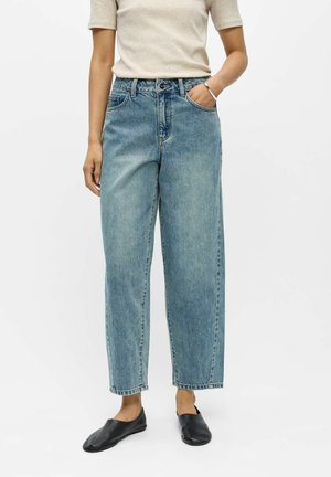 Person iført lyseblå jeans med høj talje og lige ben, beige kortærmet top og sorte slip-on sko, den ene hånd i lommen.