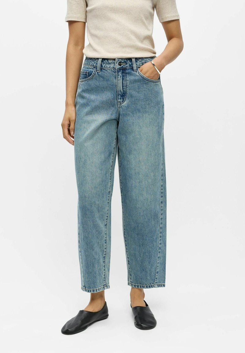 Personne portant un jean droit taille haute bleu clair, un haut beige à manches courtes et des chaussures noires sans lacets, une main dans la poche.
