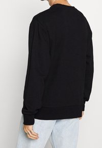 Sudadera negra de manga larga, con cuello redondo, puños y dobladillo acanalados. La tela parece suave y con textura. Combina con unos vaqueros azul claro.