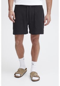 Black jersey shorts with a textured surface, featuring an elastic waistband, drawstring, and two front pockets. Worn with beige sandals.