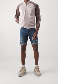 Hellrosa und braune Zip-Jacke mit drei Streifen, kombiniert mit Jeans-Shorts mit fransigen Kanten. Schuhe in neutralen Farben und weiße Socken.