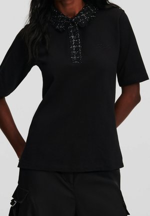 Kobieta ubrana w czarną bluzkę z krótkim rękawem, z fakturowanym kołnierzem i listwą z guzikami, w połączeniu z czarnymi spodniami bojówkami.