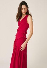 Mujer con cabello largo y oscuro, lleva un vestido rojo sin mangas con volantes, está de pie contra un fondo claro y liso, mirando hacia abajo.