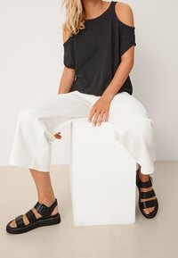 Femme portant un haut noir à épaules dénudées, un pantalon large blanc et des sandales noires à lanières, assise sur un cube blanc devant un fond neutre.