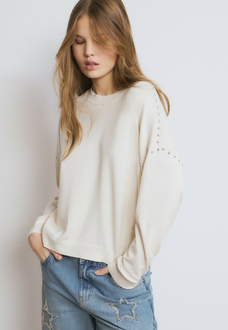 Sudadera color crema con cuello redondo y puños acanalados. Presenta tachuelas decorativas plateadas a lo largo de los hombros. Se lleva con vaqueros azules desgastados.