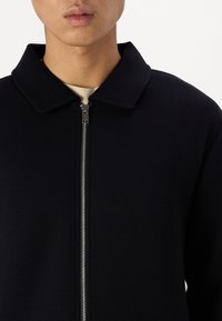 Veste noire zippée avec un design à col, dotée d'une fermeture éclair en métal et d'une texture lisse. Silhouette simple et moderne.