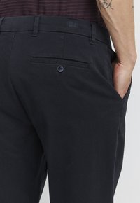 Pantalon en tissu noir avec un design épuré, une poche arrière avec fermeture à bouton et une texture subtile.