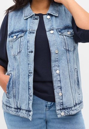 Mujer que lleva una chaqueta vaquera sin mangas de color azul claro sobre una camisa azul marino, con las manos en los bolsillos de la chaqueta, mostrada desde el cuello hasta las caderas.