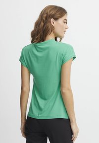 Tricou verde cu mânecă scurtă din bumbac moale, cu guler rotund și o croială lejeră, văzut din spate. Sunt purtate pantaloni negri.