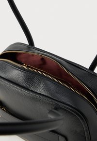 Bolso de mano de cuero negro con superficie texturizada, con una cremallera dorada y un forro interior burdeos. Diseño compacto con correas para el hombro.