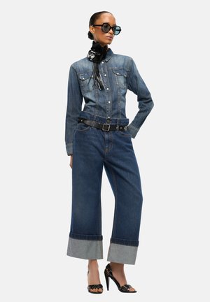 Femme portant une veste en denim foncé et un jean large avec revers, ceinture noire, accessoire de cou en forme de fleur noire, lunettes de soleil et sandales à talons hauts noires.