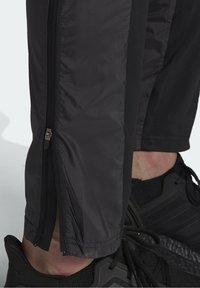 Svarta träningsbyxor med dragkedjeöppning på sidan, tillverkade av lätt material. Har en slät yta och är kombinerade med svarta sneakers.