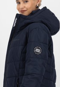Marine blauwe pufferjas met capuchon, voorzien van een ritssluiting en een logo-opdruk op de linker mouw. Het materiaal lijkt glad en licht gewatteerd.