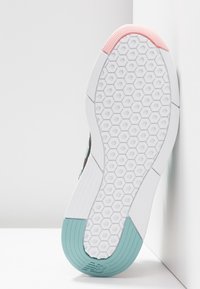 Semelle de chaussure avec motif de crampons hexagonaux, base blanche, grip rose à l'avant-pied et grip turquoise au talon, appuyée contre un mur blanc.