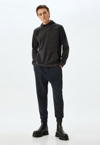 Mörkgrå huvtröja med ficka fram, i kombination med matchande mörka joggingsbyxor och kraftiga svarta stövlar. Slät textur, avslappnad passform.