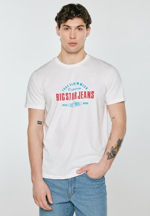 Młody mężczyzna z kręconymi włosami, ubrany w białą koszulkę Big Star Jeans i niebieskie jeansy, z tatuażami na lewym przedramieniu i prawej ręce.