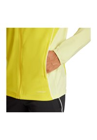 Gelbe Sportjacke mit glatter Textur, ausgestattet mit einem Reißverschluss, seitlichen Taschen und kontrastierenden weißen Akzenten. "AEROREADY" Logo abgebildet.