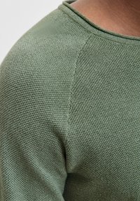 Close-up of a person wearing a textured, green knitted sweater with visible shoulder seam and round neckline.