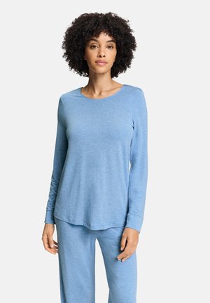 Donna con capelli ricci che indossa una maglia a maniche lunghe azzurro chiaro e pantaloni abbinati, in piedi davanti a uno sfondo chiaro e uniforme.